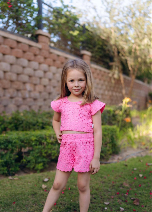 A child wearing a pink outfit consisting of a top and shorts, standing on grass with a garden and a brick wall in the background.