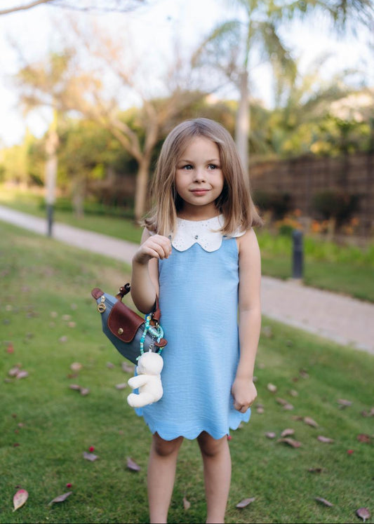 A young girl standing on grass wearing a blue sleeveless dress with a white collar, holding a toy.