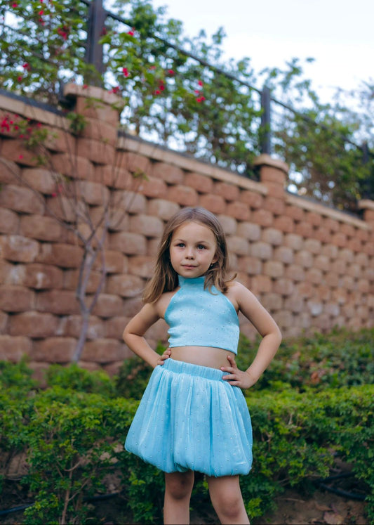 A young girl wearing a blue sleeveless top and skirt standing near a brick wall with vines and red berries on the wall in the background.