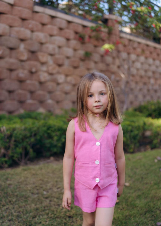 Young girl in a pink outfit standing outdoors near a stone wall and greenery.