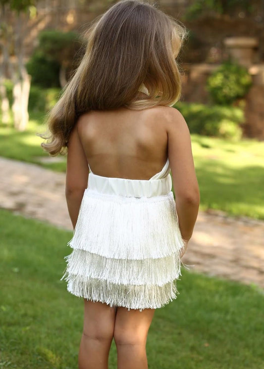 Child in a white fringe dress walking outdoors on a path surrounded by greenery.