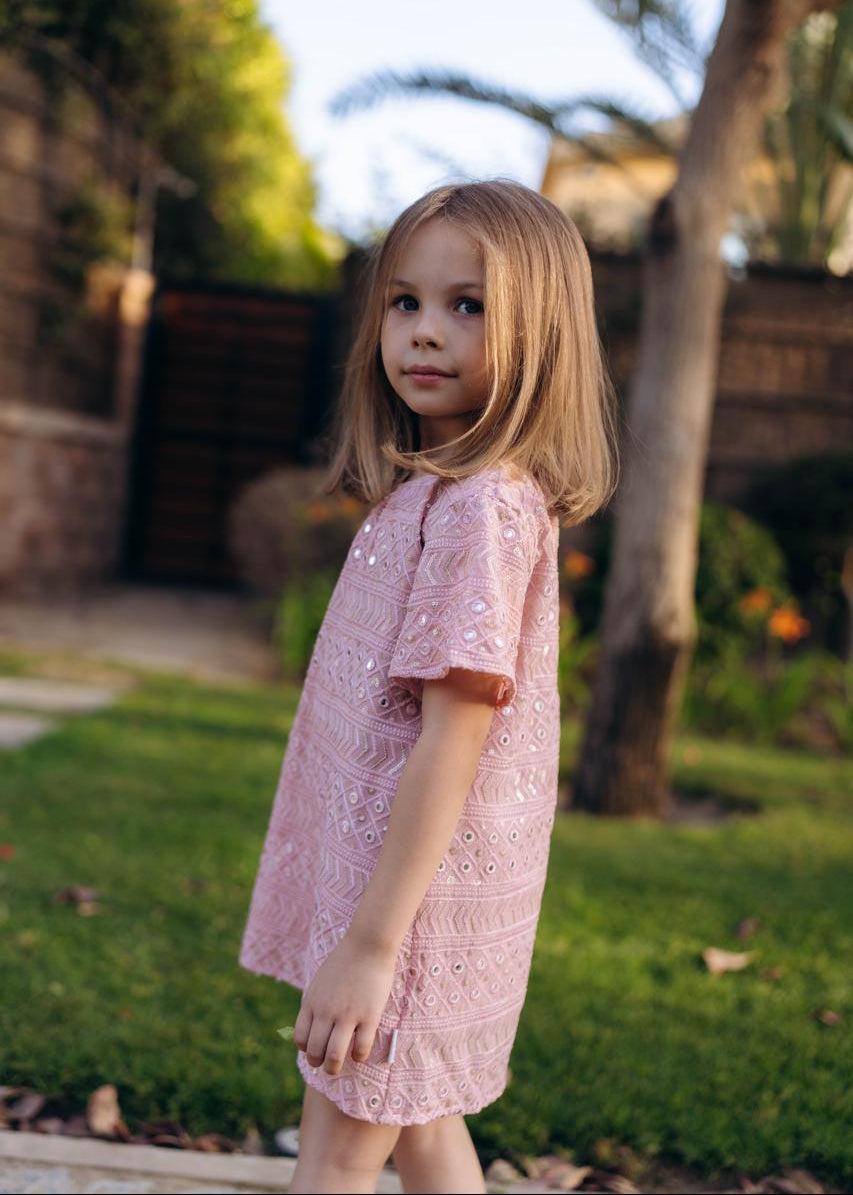 Young girl in a pink lace dress standing outdoors with a garden background.