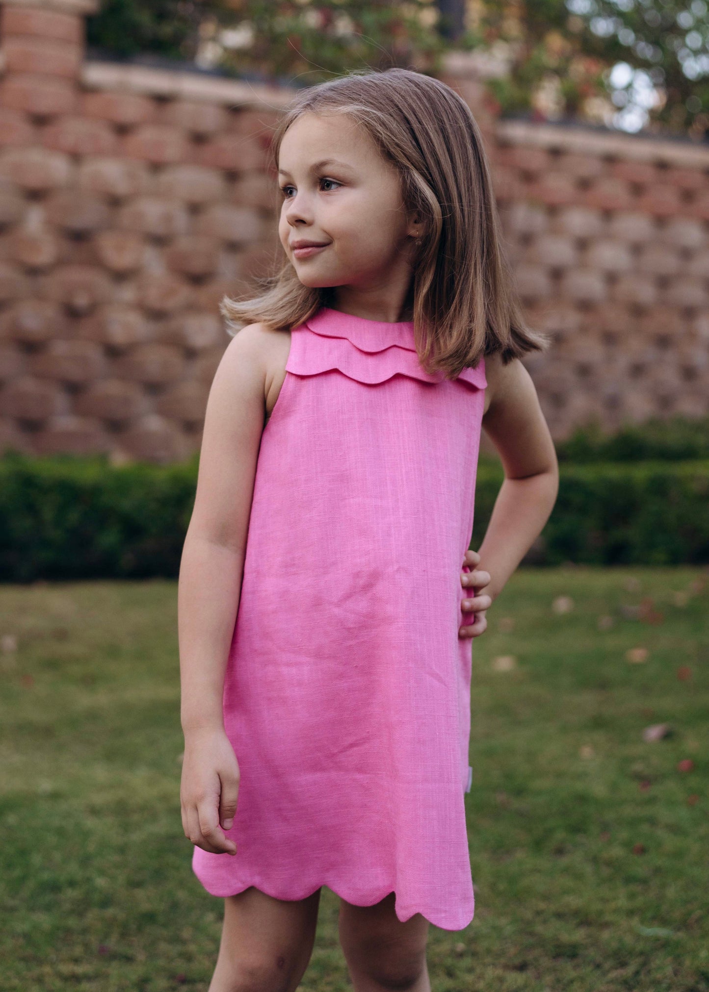 Young girl in a pink dress standing outdoors near a brick wall and hedges.