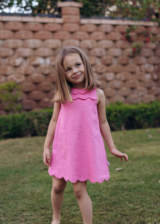 A young child standing on grass against a stone wall backdrop, wearing a pink sleeveless knee-length dress with a scalloped edge.