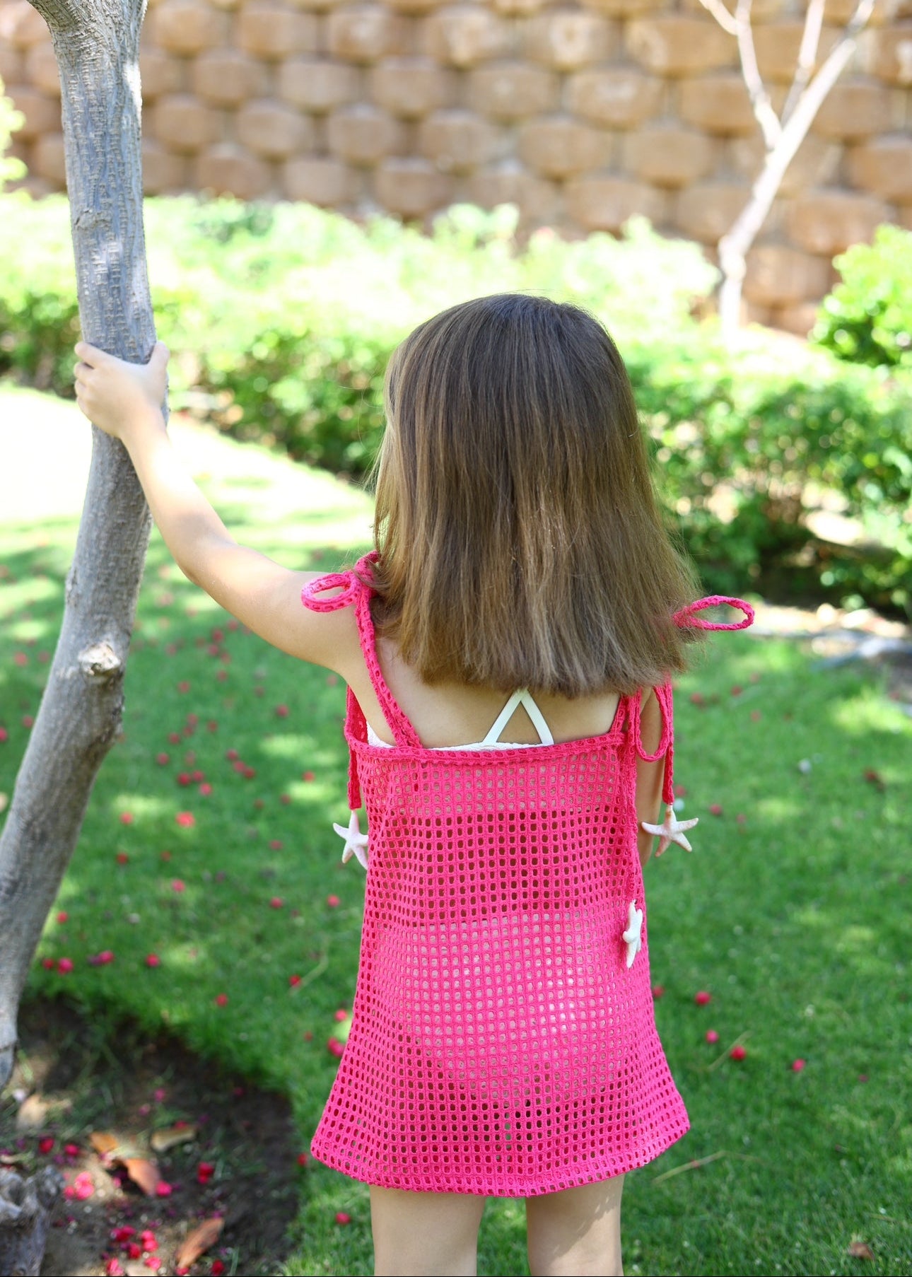 Child in a pink dress standing outdoors with a tree and garden in the background