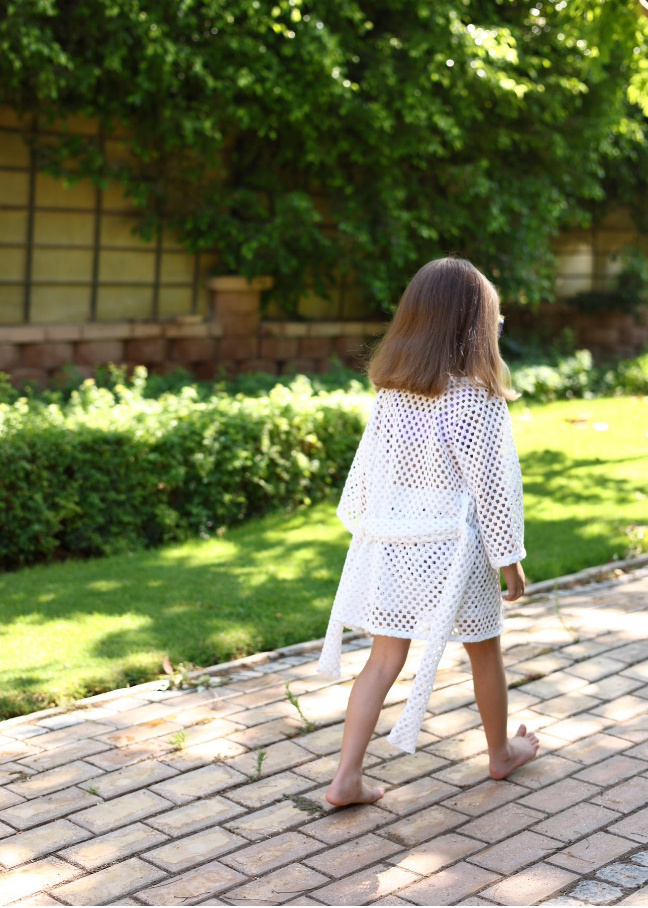 Person wearing a white crochet cover-up walking on a paved path with greenery in the background