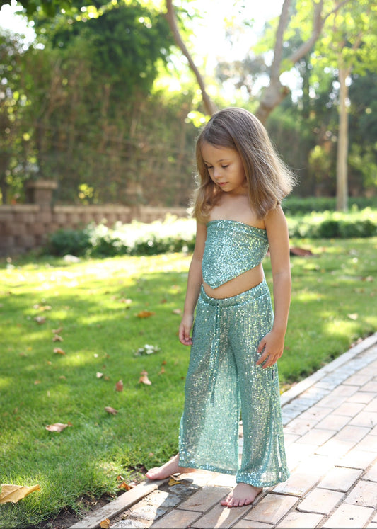 Young girl in a sparkly green dress standing on a paved path with grass and trees in the background