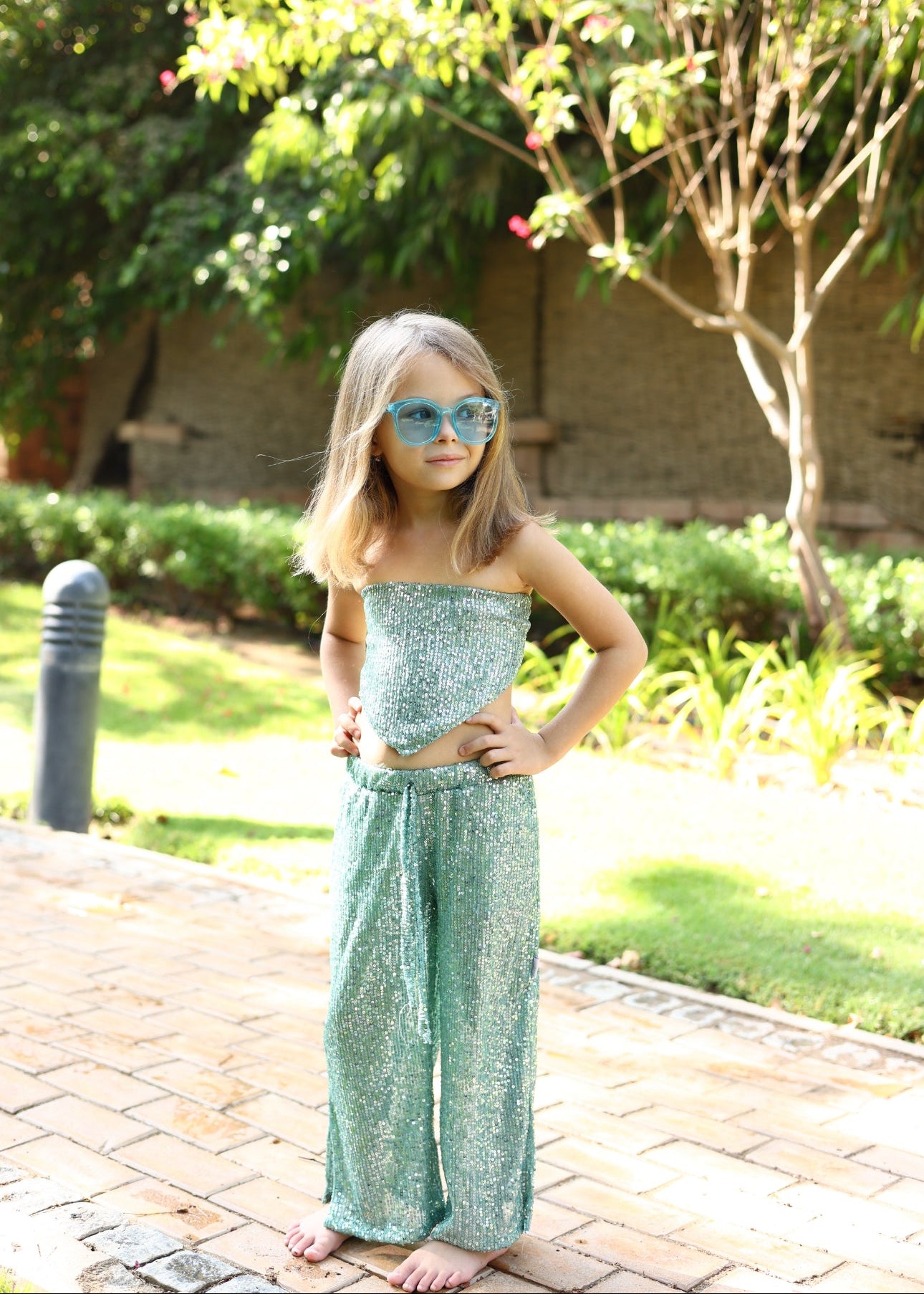 Young girl in a green dress and sunglasses standing outdoors on a paved path with trees in the background.