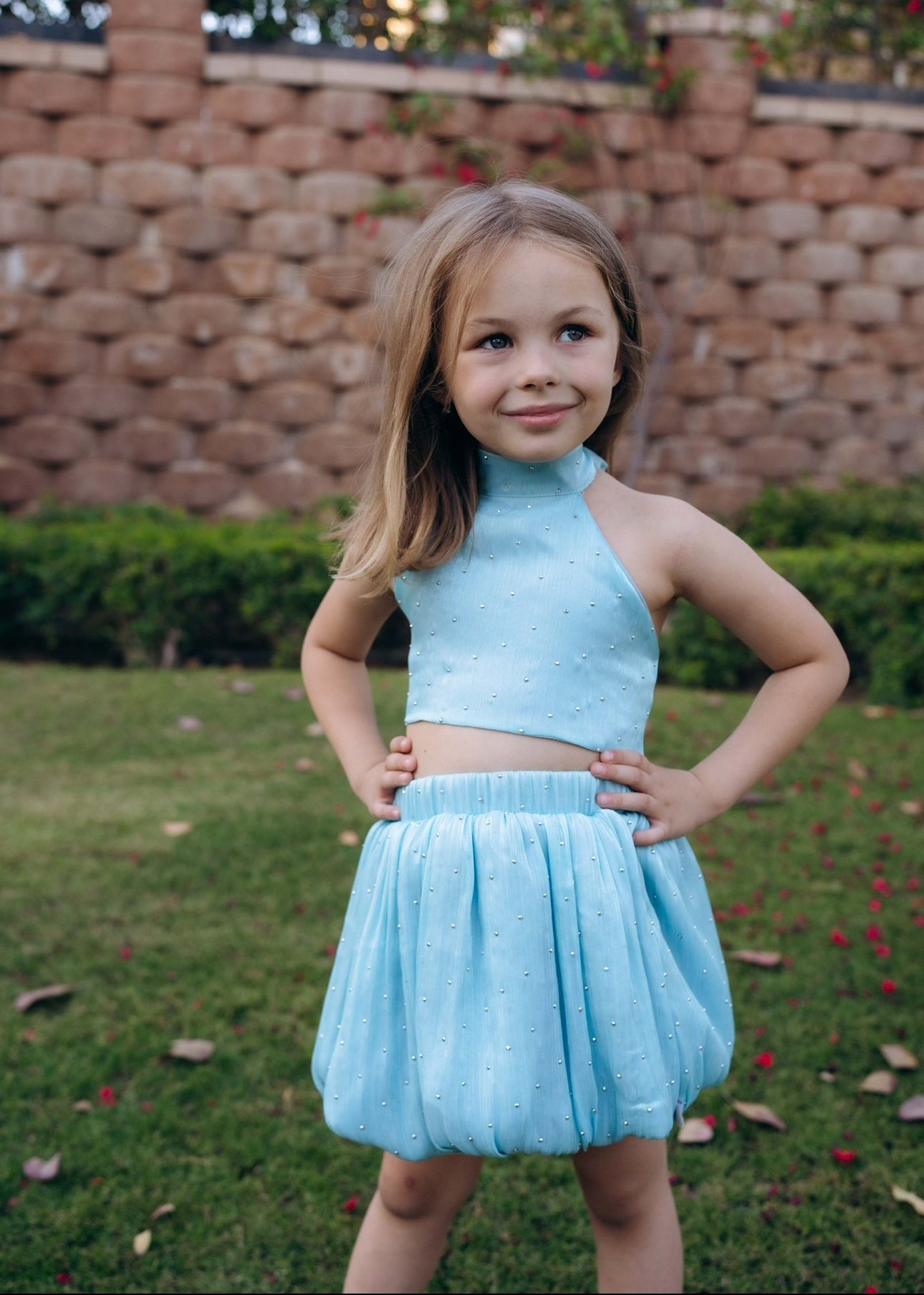 Young girl in a light blue outfit standing outdoors with a brick wall and greenery in the background