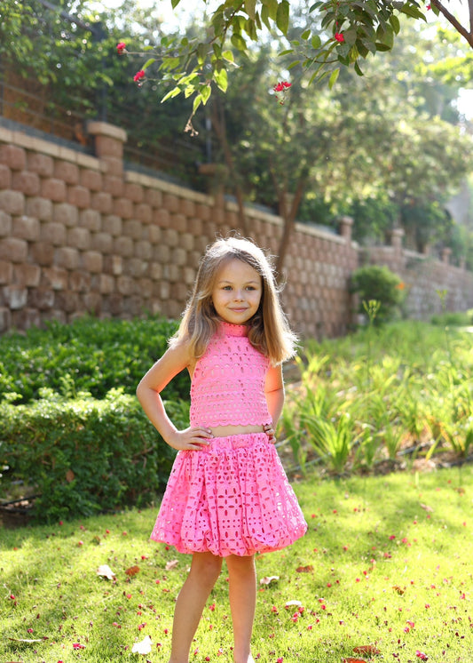 Young girl in a pink dress standing outdoors with greenery and a stone wall in the background