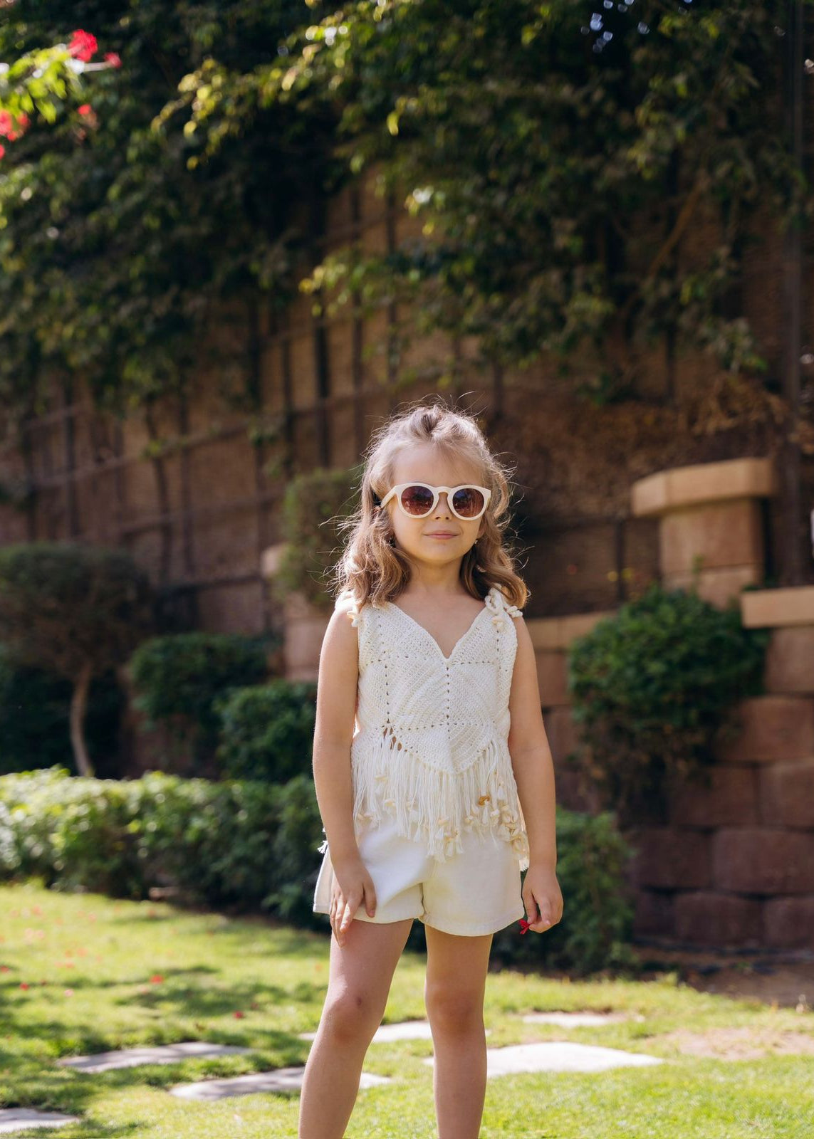Young girl in a white dress and sunglasses standing in a garden.