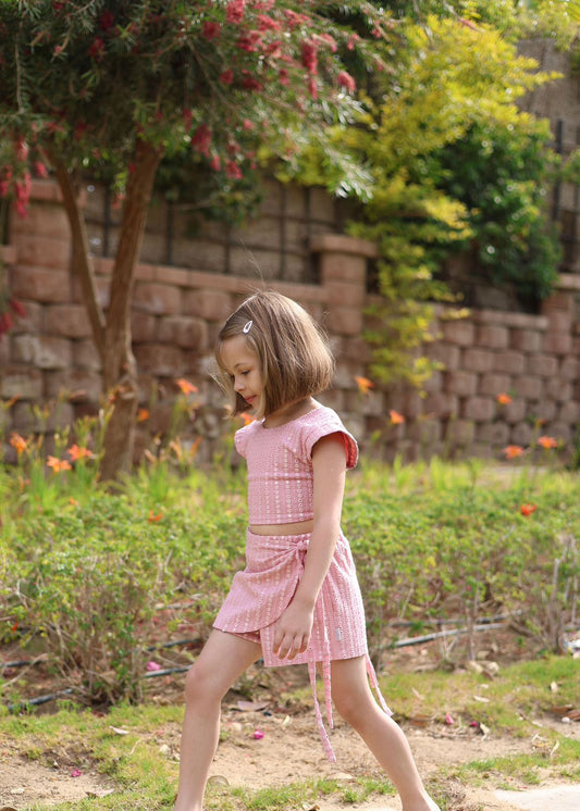 Young girl in a pink dress standing in a garden with flowers and a stone wall in the background.