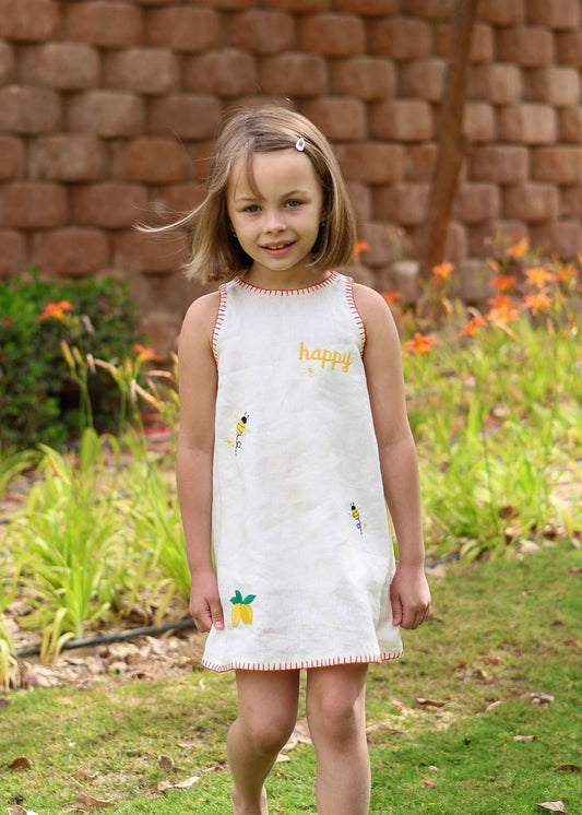 Young girl in a white dress with colorful designs standing outdoors in front of a brick wall and flowers.