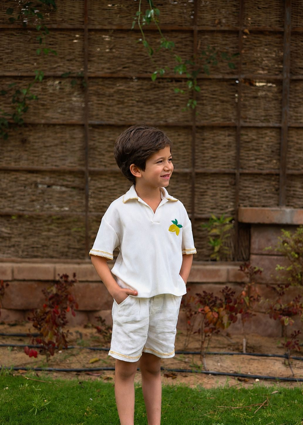 Young boy in a white outfit standing in front of a wooden fence.