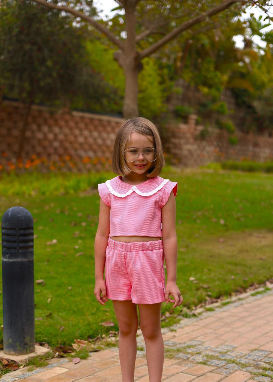Young girl in a pink outfit standing outdoors on a grassy area with trees in the background.