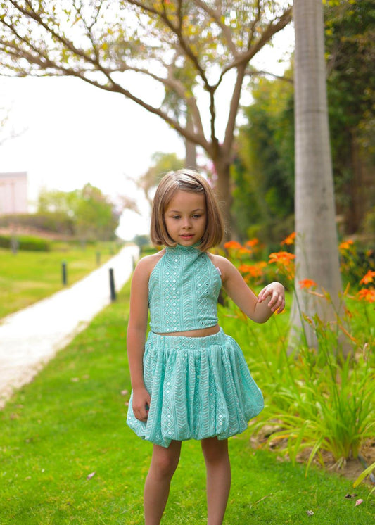 Young girl in a turquoise dress standing in a garden with flowers and trees.