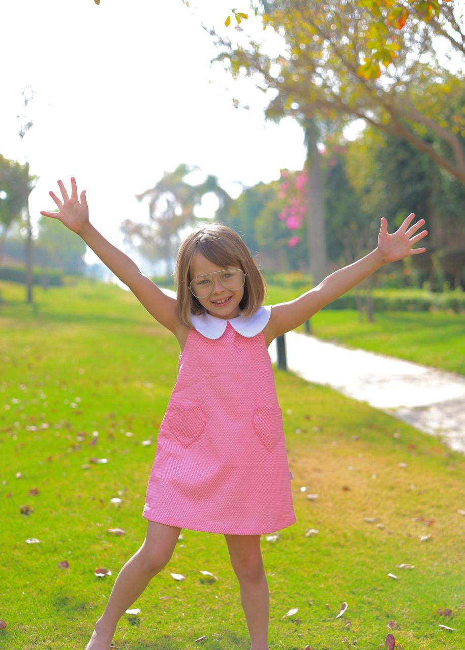 Young girl in a pink dress with a white collar standing in a park with trees and grass.