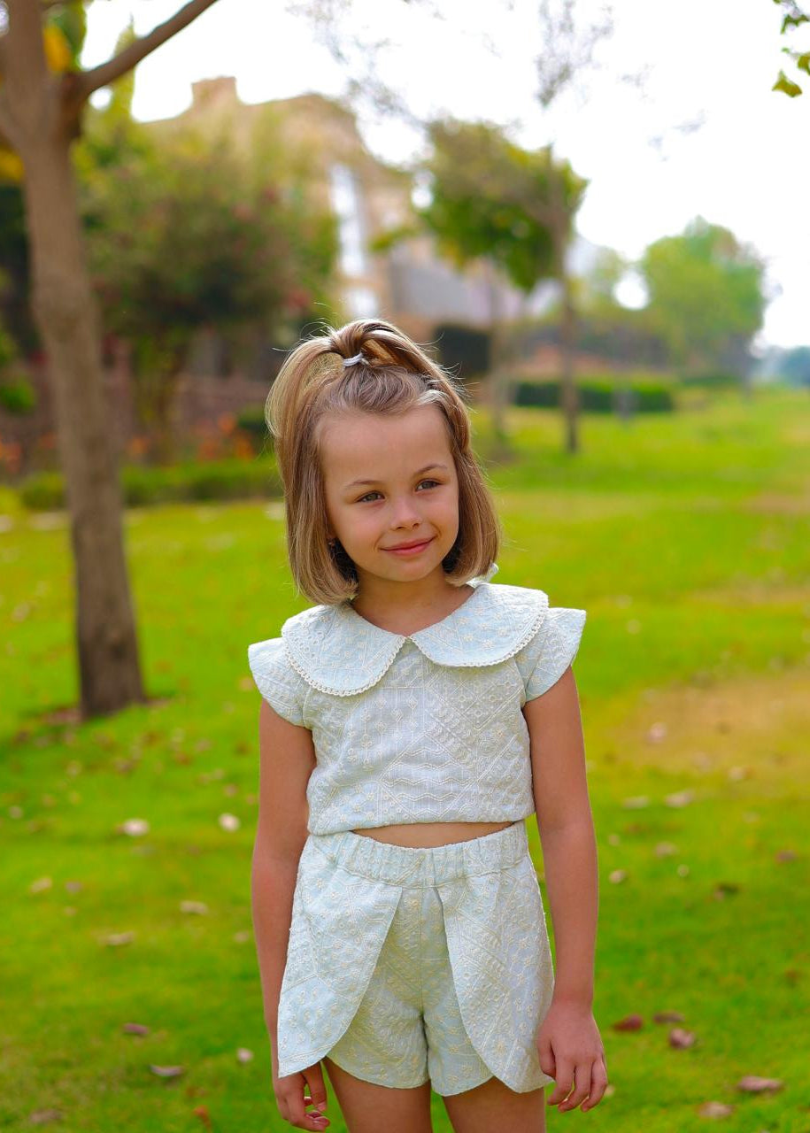 Young girl in a white outfit standing in a park with green grass and trees.