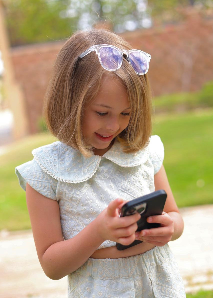 Young girl wearing sunglasses and a white dress, looking at a phone outdoors.